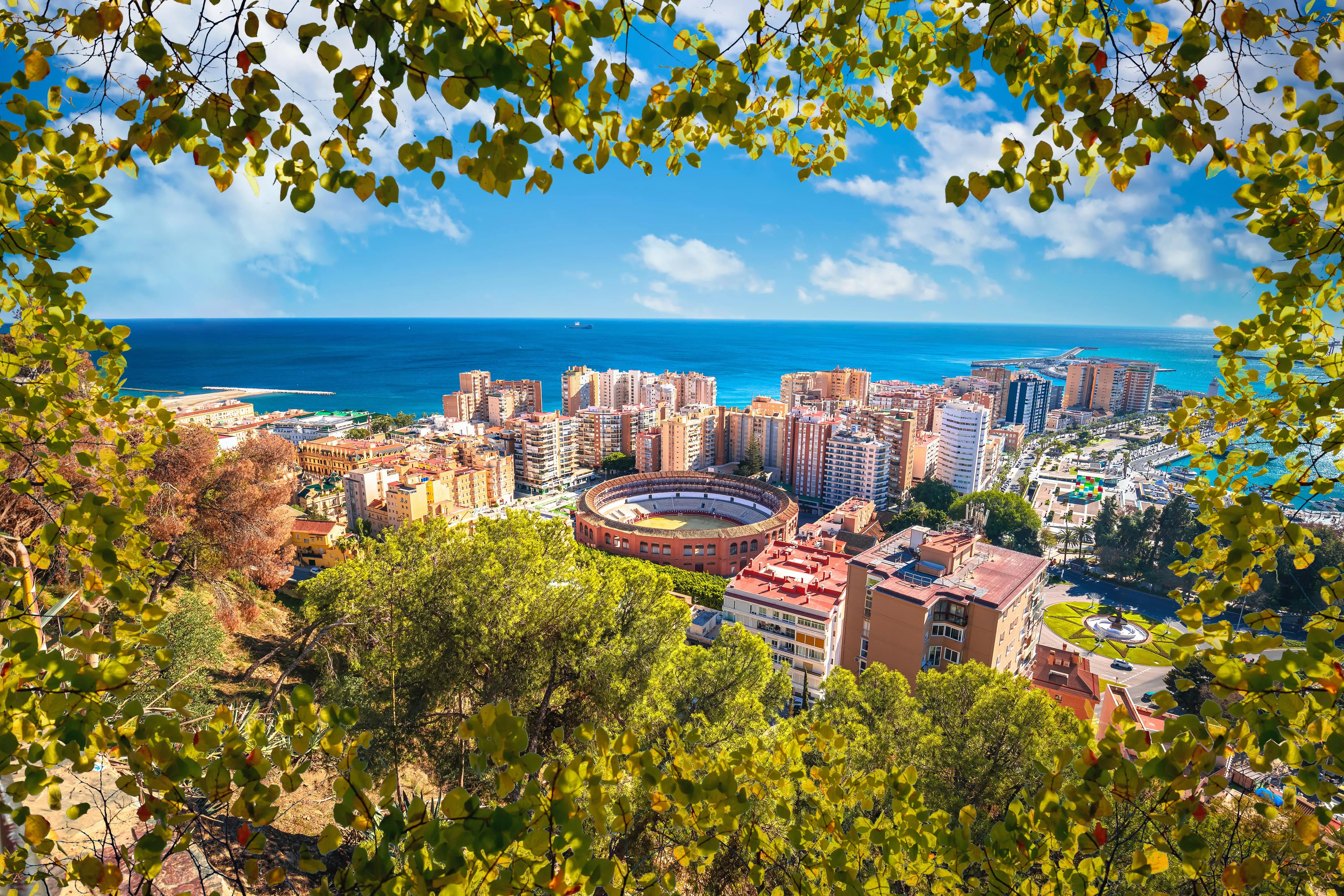 Panoramisch uitzicht op Málaga met de haven, arena en zee, omlijst door groene bladeren en een blauwe lucht.