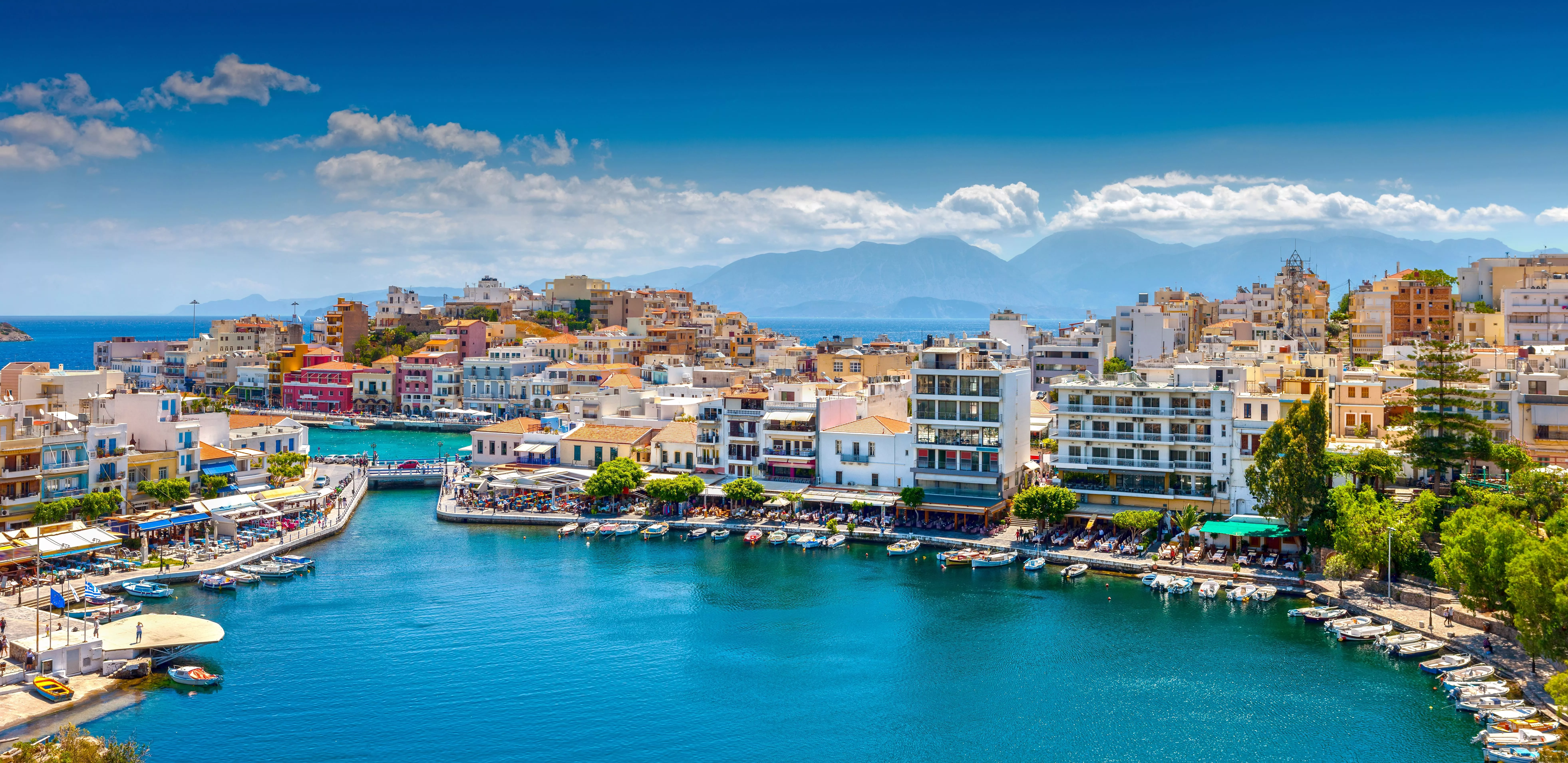 Kleurrijke huizen en bootjes rond de haven van Agios Nikolaos op Kreta, met bergen en zee op de achtergrond.