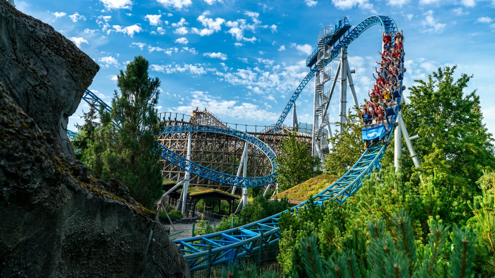 Blauwe achtbaan in een pretpark, met een kar vol juichende bezoekers in een scherpe bocht.